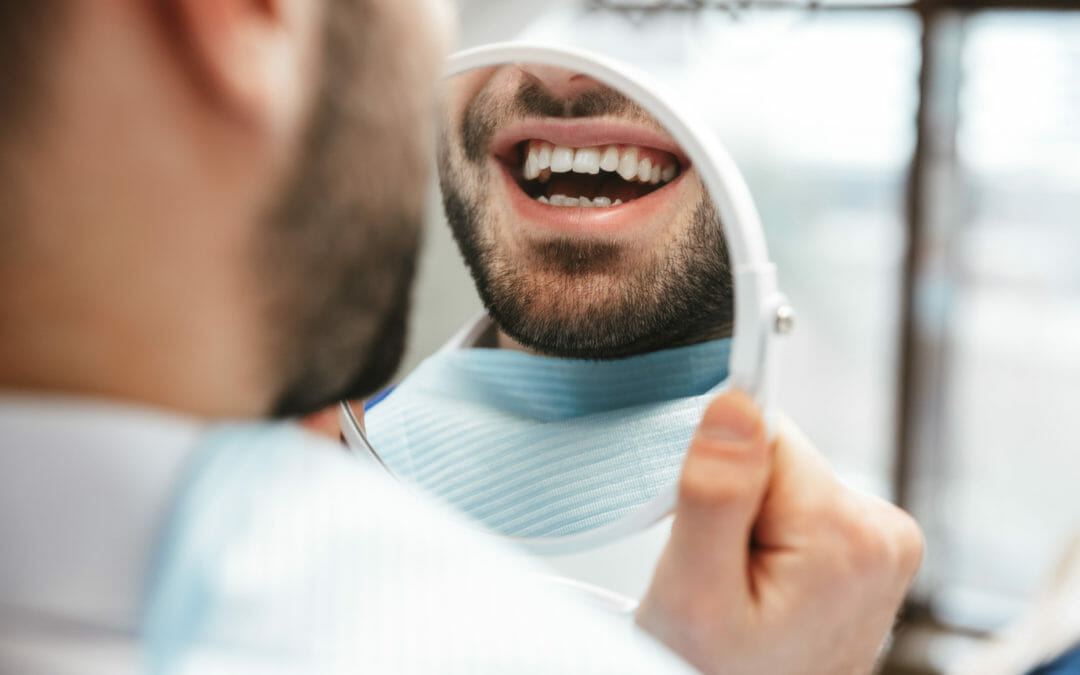 man smiling and looking at his teeth in handheld mirror